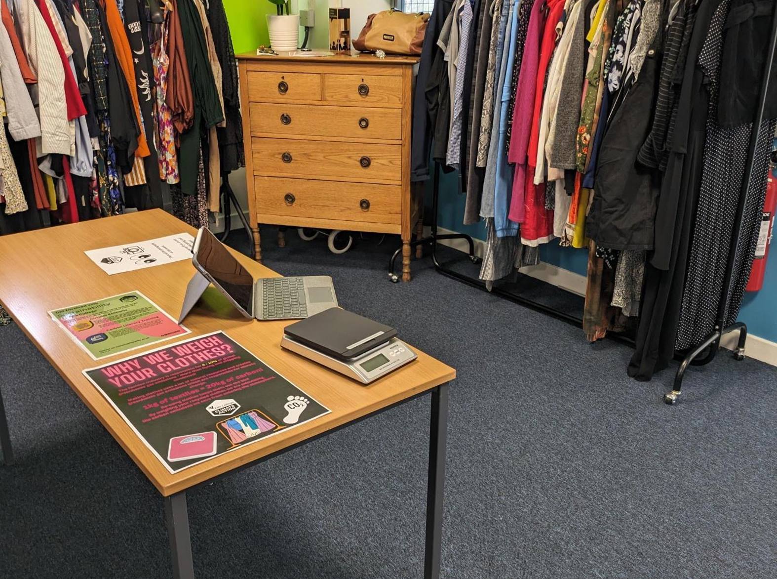 room with two clothes rails, clothes on the clothes rails, set of wooden drawers and a table